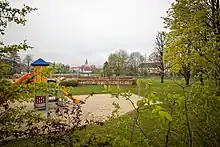 Fotografie eines Geländes mit Sandgrube, in der ein buter Plastikrutschenturm steht. Äste einer Hecke und von Bäumen mit jungem Grün ragen von beiden Seiten und von unten in das Bild