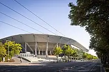 Foto eines weißen Stadiongebäudes über eine Straße mit Trambahnschienen und -leitungen hinweg.
