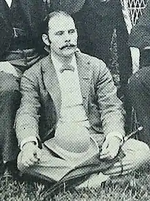 Edward John Cameron, 1885. Schwarz-Weiß-Bild von einem Mann mit dünnem Schnurrbart im Schneidersitz.