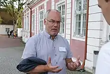 Frontale Farbfotografie von einem Mann, der zu einer Person am rechten Bildrand spricht. Mit seinen Händen gestikuliert er angespannt vor seiner Brust. Er hat lichte, graue Haare und trägt eine Brille und einen Dreitagebart. An seinem kurzärmeligen, blauen Hemd steckt rechts ein Namensschild und er hat sein dunkelblaues Jackett über seinen rechten Arm gelegt.