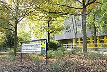  Eingang zum Berufsförderungswerk Düren, ein Schild mit Firmen-Beschriftung und Logo steht unter einem Baum im Vorgarten zum BFW