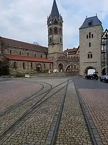 Ein städtischer Platz. Im Hintergrund stehen Gebäude, ein Turm und eine Tordurchfahrt. Im Straßenpflaster befinden sich zwei schmalspurige Gleise, die mit zwei Weichen verbunden sind