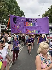 In einer Straße marschieren Personen verschiedenen Alters in einem Umzug. Vorne stehen mehrere Frauen, von denen zwei ein lila Stoffplakat hochhalten. Dieses ziert die mit verschiedenfarbigen Buchstaben verfasste, englische Aufschrift „Jeder weiß, dass die schwarze Lesbe Stormé die Auslöserin von Stonewall war“. In der linken oberen Ecke ist ein Schwarz-weiß-Porträt von Stormé.