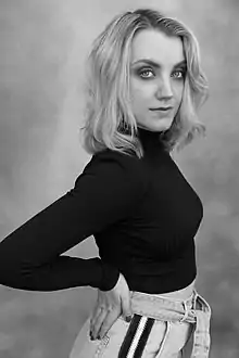Schwarzweißfotografie von einer jungen Frau im Halbprofil. Sie blickt selbstbewusst in die Kamera und hat ihre rechte Hand am oberen Gesäß platziert, das seitlich zu sehen ist. Sie trägt helles, schulterlanges Haar, das leicht gewellt ist. Ihr schwarzer Rollkragenpullover ist gecroppt und lässt ein wenig den Bauch erblicken, der zur hellen Jeans übergeht.