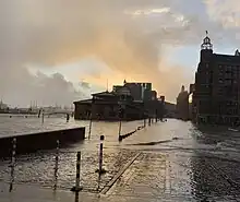 Hamburger Fischmarkt unter Wasser während der Sturmflut 2020 am 10. Februar, verursacht durch Orkan Sabine. Der Himmel über der im Wasser stehenden Fischmarkthalle ist in ein fahl gelblich-oranges Licht getaucht, die spätnachmittägliche Sonne scheint durch ein Wolkenloch in die ansonsten graue-düsteren Wolken. Einige vom Wind angetriebene kleine Wellen spülen über das nasse Kopfsteinpflaster der Straße, Straßenlaternen und die Bushaltestelle vor dem länglichen Gebäude am Ufer stehen im Wasser, das Licht spiegelt sich in Glasfenstern einer ansonsten dunklen Fassade auf der gegenüberliegenden Straßenseite rechts und auf der Wasseroberfläche. Der Landungssteg links im Bild ist teils von Elbwasser überspült, dahinter Hafenkräne in der Ferne und einige dreiflügelige Windkraftanlagen.