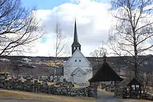Foto einer weißen Holzkirche mit dunklem Kirchturm. Die Kirche ist von einem Friedhof umgeben, der wiederum von einer Steinmauer umgeben ist. Im Hintergrund eine hügelige Landschaft.