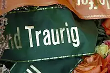 Das Wort „Traurig“ in goldener Schrift auf einem dunkelgrünen Stoff, am Rande des Bilds sind weitere Trauerschleifen in anderer Farbe und Zweige und Blumen zu sehen.