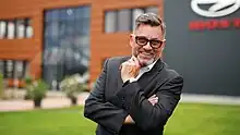ein freudlos grinsender bärtiger Mann mit Brille in einem Anzug