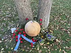 Die Gedenkkugel auf einer herbstlichen, mit Blättern bedeckte Wiese. Die Gedenkkugel ist mit roten Nelken und Rosen geschmückt und links im Bild ist ein Kranz mit blauen Schleifen zu sehen.