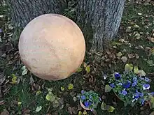 Eine beige Tonkugel liegt auf einer Wiese bei einem Baumstamm. Rechts im Bildrand sind Schalen mit Veilchen zu sehen.