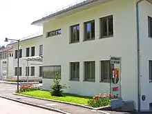 Gebäude mit heller Fassade, davor auf einer Glasstele das Wappen der Gemeinde, darüber die Aufschrift "Gemeindeamt St. Gotthard", darunter "Hort" (für die Kinderbetreuungsstätte im Untergeschoß des Gebäudes)