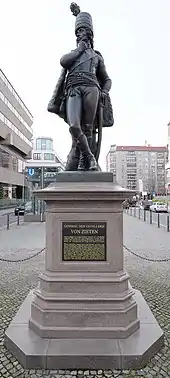 Farbige Untersicht auf eine Männerfigur mit Uniform, der seine Beine überkreuzt und sich mit der linken Hand am Dolch abstützt. Mit der rechten Hand reibt er an seinem Kinn und blickt nach links. Die Figur steht auf einem Sockel mit der Aufschrift "General der Cavallerie von Zieten".