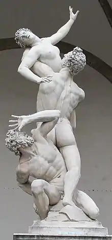 Frontale Farbfotografie in der Untersicht von drei nackten Marmorfiguren mit Lockenfrisuren. Der untere Mann hockt zwischen den Beinen des anderen Mannes auf einem Stein und blickt mit abwehrender Hand vor seinem Gesicht erschrocken nach oben. Der mittlere Mann steht mit dem Rücken zum Betrachter und hebt eine Frau über Brusthöhe. Dabei stützt er ihren Kopf mit seinem linken Arm. Die Frau hebt ihren linken Arm nach oben und stützt sich mit ihrer rechten Hand am Mann ab.