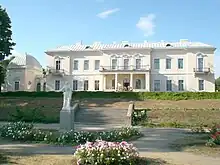 Frontale Farbfotografie eines zweigeschossigen neobarocken Gebäudes. Das beige Gebäude ist mit weißen Ornamenten und Giebelfeldern verziert. Der Mittelteil hat Säulen und eine Terrasse sowie im Obergeschoss einen Balkon. Links ist ein Nebengebäude mit einer Kuppel. Der Garten mit Blumenbeeten und einer weißen Skulptur ist über eine kurze Treppe zu erreichen.