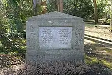 Rechteckiger Grabstein mit aufgesetztem Dreieck, alles aus grauem Stein. Mittig eine heller Steinplatte mit Lebensdaten von vier Personen, unten rechts die von Helga Martin. Vor dem Grabstein wächst Erika, im Hintergrund sieht man Bäume und einen bemoosten Pfad.