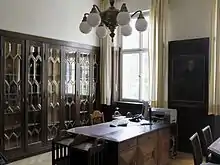 Schwere dunkle Möbel: Ein Schreibtisch und mehrere Aktenschränke mit Glasfronten, an der Wand ein Porträt von Prinzregent Luitpold. Das Büro wird weiterhin genutzt, deshalb stehen auf dem alten Schreibtisch ein Computer und ein Telefon, an der Wand ein Drucker