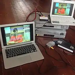 Ein Laptop mit den Aufnahmen des Spiels steht auf zwei VHS-Rekordern. Drähte verbinden sie zu einem naheliegenden Laptop, connect them to another laptop nearby, mit einer VHS-Kartusche auf der rechten Seite.