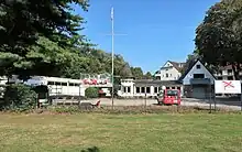 Hinter einem Rasenstück und einem Zaun liegt ein Parkplatz. Dahinter sind ein Bootsjaus und ein Vereinslokal zu erkennen.