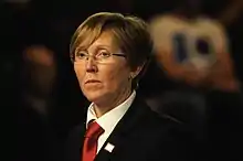 Das Bild zeigt Hilde Moens in einer Naheinstellung. Sie steht aufrecht und schaut mit ernstem Blick links am Betrachter vorbei. Sie hat halblanges, dunkelblondes Haar und trägt eine Brille, kleine, slberne Ohrringe, ein weißes Oberhemd, ein schwarzes Jackett und eine rote Krawatte.