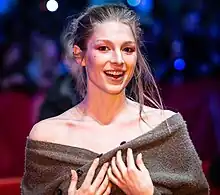 Hunter Schafer steht auf einem roten Teppich und hält ihre Hände mit gespreizten Fingern auf die Brust. Sie trägt einen aufgeknöpften, leicht heruntergezogenen dunkelbraunen Pullover, der ihre Schultern nur teilweise bedeckt, rosa Gelnägel an den Fingern sowie ihre blonden Haare strähnig und hinten über den Rücken, vorne bis zur linken Schulter. Im Hintergrund sind unscharfe Personen, ein dunkelrotes Seitenbanner und dunkelblau-weiße Lichter zu sehen.