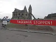 Banner an Bauzaun: Tagebaue stoppen!, Kirche und zwei Abrissbagger im Hintergrund