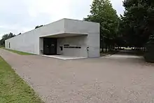 Ein flaches Gebäude aus Glas und Beton steht an der Ecke eines Feld- und eines Alleeweges