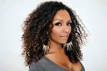 Porträtfoto von Janet Mock vor weißem Hintergrund. Sie trägt ein graues Oberteil, Wimperntusche, goldfarbene, große Ohrringe mit in der Mitte runterhängenden Spitzen und ihre braunen Haare als Locken.
