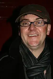 Joe Pasquale steht vor einer dunkelroten Wand. Er trägt einen schwarzen Kapuzenpullover, ein graues Hemd, einen schwarz-grau-blau gestreiften Schal, eine schwarze, ovale Brille und eine dunkelbraune Schirmmütze mit einer kleinen, roten Boss-Aufschrift, einem kleinen, roten Quadrat und einer roten gestrichelten Linie auf dem Schirm.