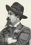 Jorge Collaço trägt einen großen Hut und hat einen Oberlippenbart und längeren Bartwuchs am Kinn. Die schwarz-weiß-Fotografie zeigt ihn mit einem Anzug gekleidet und verschränkten Armen.
