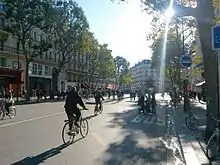 Seitliche Farbfotografie einer Allee mit Fahrradfahrern und Fußgängern auf der Straße. An den Seiten sind alte Häuserfassaden mit Geschäften. Rechts ist eine Busfahrbahn mit den dazugehörigen Hinweisschildern auf dem Bürgersteig.