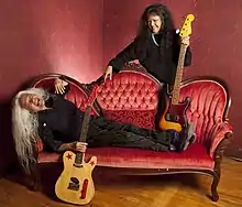 June Millington mit weißen langen Haaren liegt lachend auf einem roten Sofa, mit einer Gitarre in der Hand. Jean mit schwarzen langen Haaren steht hinter dem Sofa, hat einen elektrischen Bass und lacht June an.