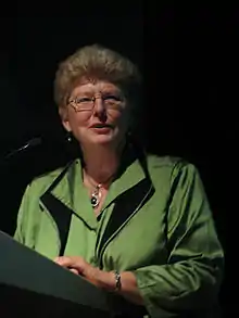 Eine Frau steht an einem Rednerpult und spricht in ein Mikrofon. Sie hat kurze blonde Haare und trägt eine grüne Jacke.