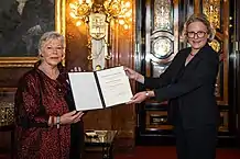 Katrin Rohde hält eine Urkunde für das Bundesverdienstkreuz in der Hand. Neben ihr steht Staatsrätin Almut Möller und übergibt es ihr.