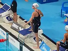 Stewart trägt einen dunkelblauen Badeanzug und eine Schwimmbrille. Auf der weißen Badekappe ist die US-Flagge und ihr Nachname zu sehen. Stewart hat das rechte Bein bereits auf dem Startblock, das linke Bein steht noch auf dem Beckenrand.