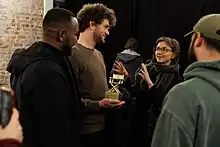 Eine Gruppe Menschen vor einer Bühne. Ein junger Mann hält eine Filmpreis in Form eines goldenen Regiestuhls in seinen Händen