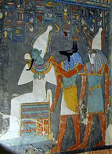 Gemaltes Relief eines sitzenden Mannes mit grüner Haut und engen Gewändern, eines Mannes mit dem Kopf eines Schakals und eines Mannes mit dem Kopf eines Falken