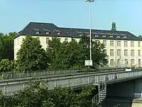 Eines der ursprünglichen Magazingebäude. Ansicht vom Nordostrand des Langemarckplatzes außerhalb der Liegenschaft aus, Blickrichtung: südwest, die auf dem Bild sichtbare lange Seite des Gebäudes zeigt Richtung Nordosten; die über die Brücke im Vordergrund führende Straße ist die B 416 Mayener Straße, das silberne Auto fährt Richtung Westen, unter der Brücke hindurch führt die B 9. (Die unten dargestellte Photopostkarte zeigt dieses Gebäude vor Kriegsbeschädigung und Wiederaufbau).