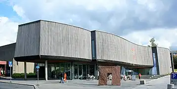 Foto eines modernen Gebäudes. Das unterste Stockwerk hat eine Glasfassade, die darüberliegende Etage steht stark hervor und ist nahezu fensterlos.