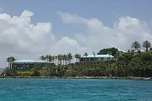 paradiesische Insel, sehr schönes blaues Wasser vorn, Wolken im Hintergrund, zwei flache Häuser und Palmen und anderer Grünwuchs