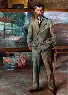  Ein Gemälde mit Eichfeld in seinem Atelier im Vollporträt. Er steht vor einigen seiner Bilder und trägt einen grauen Anzug. Das Haar ist dunkel und an den Schläfen grau, er trägt zudem einen gestutzten Vollbart.