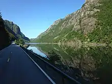 Bild von einer Straße, die entlang eines Gewässers führt. Links und rechts erheben sich teils begrünte, teils felsige Berge.