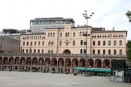 Foto eines Gebäudes mit verzierter Fassade und einer Uhr darin, davor ein Platz