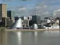 Mittig im Bild befindet sich das Science Center umgeben von einer Skyline aus Hochhäusern. Vordergründig ist Wasser zu sehen.