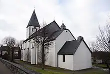 Von links nach rechts: Kirchturm, Hauptschiff mit Stützpfeilern, Chor (niedriger) und Sakristei (nochmals niedriger) der katholischen Pfarrkirche St. Margaretha Madfeld; alle Wände weiß gestrichen, schieferbedeckte Dächer. Fensterlose Rückansicht der Kirche mit Kirchhof, darauf laubfreie Bäume.