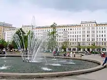 Frontale Farbfotografie eines mehrstöckigen, weißen Gebäudes, das über den ganzen Hintergrund geht. Weiter links und rechts von der Fassade sind eckige Vorbauten. Die vielen Fenster sind schmal und das Dach hat ein blau-goldenes Geländer. Im Vordergrund steht ein Springbrunnen mit Menschen.