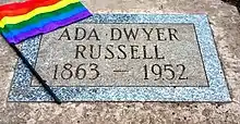 Auf einer grauen Steinplatte mit silbrig-blauer Umrandung ist eine Gravur zu erkennen: Ada Dwyer Russel 1863–1952. Daneben liegt eine Regenbogenflagge.