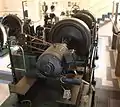 Maschinensatz mit zwei Turbinen (Hintergrund) auf einer Welle, Regelungseinrichtung mit Flachriemen angetrieben und Generator im Vordergrund