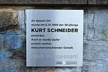 Silberne Gedenktafel mit schwarzer Schrift befestigt auf einer Steinmauer. Darauf steht: "An diesem Ort wurde am 6.10.1999 der 38-jährige Kurt Schneider ermordet. Auch er wurde Opfer extrem rechter, menschenverachtender Gewalt. Kein Vergessen!"