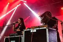 Beide Frauen sind auf der in rotes Licht getauchten Bühne zu sehen Hélène de Thoury steht am Synthesizer mit wehenden Haaren. Die hinter einem Synthesizer stehende Amandine Stioui hält sich das Mikrophon vor den Mund und singt.