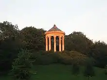 Farbfotografie in der Untersicht eines Rundtempels mit Säulen und einer goldenen Kugel als Spitze. Der Tempel steht auf einem Hügel mit Büschen und ist von Bäumen umgeben.